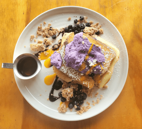 A plate of pancakes topped with purple whipped cream, blueberries, and drizzled syrup, surrounded by granola.
