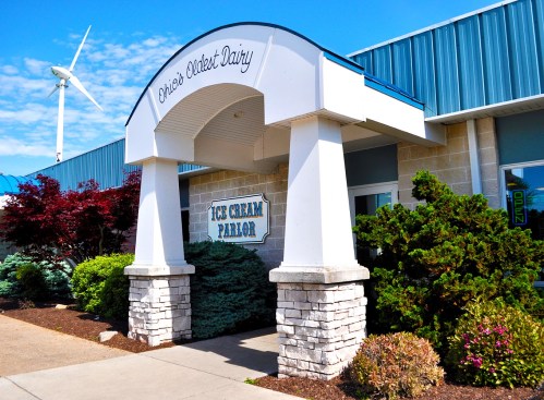Entrance of an ice cream parlor with a sign reading "Ohio's Oldest Dairy," surrounded by greenery and a wind turbine in the background.