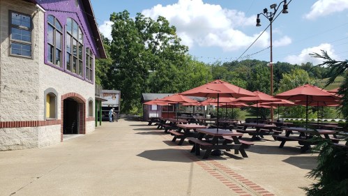 Outdoor seating area with picnic tables and umbrellas, next to a building with large windows and greenery in the background.