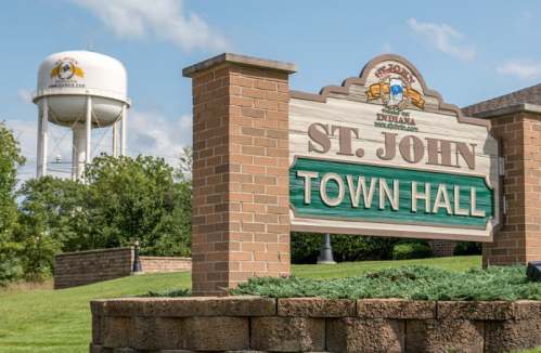 Sign for St. John Town Hall with a water tower in the background under a blue sky.