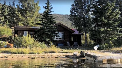 A rustic cabin surrounded by trees, with a dock extending into a calm lake and mountains in the background.