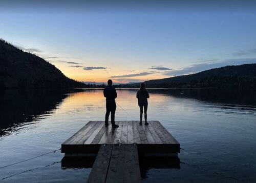 Two people stand on a wooden dock by a calm lake at sunset, surrounded by mountains and a colorful sky.