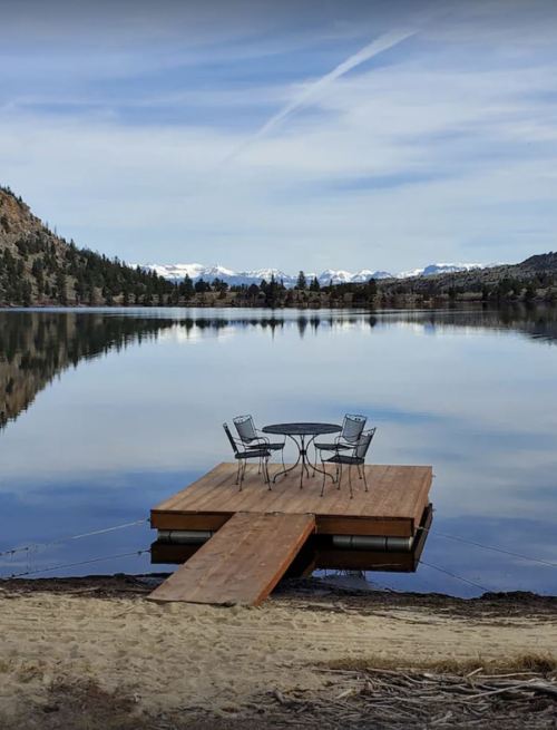 A wooden dock with a table and chairs overlooks a calm lake surrounded by mountains and trees.