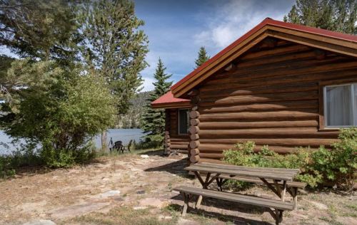 A rustic log cabin by a lake, surrounded by trees and featuring a picnic table in the foreground.