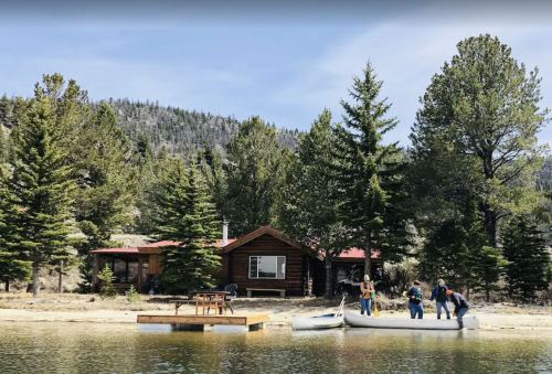A rustic cabin by a lake, surrounded by pine trees, with people preparing canoes on the shore.