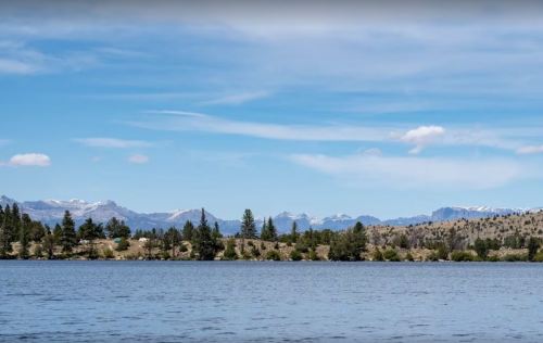 A serene lake surrounded by trees and mountains under a clear blue sky with a few clouds.