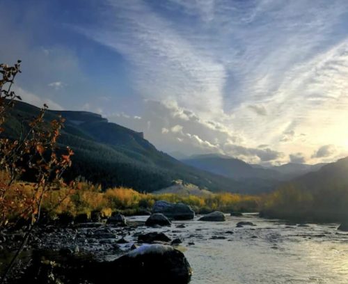 A serene river flows through a valley, surrounded by mountains and colorful autumn foliage under a cloudy sky.