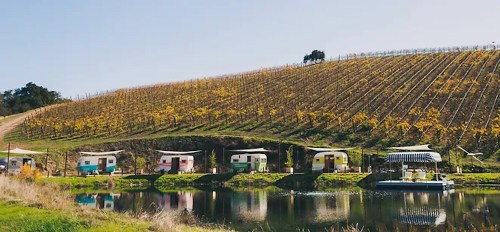Colorful vintage trailers by a serene lake, set against a backdrop of lush vineyards and a clear blue sky.