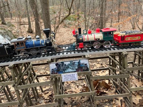 A model train set on a wooden track in a forest, with a sign displaying "Mark's Miniature World" nearby.