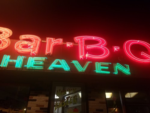 Neon sign reading "Bar-B-Q Heaven" illuminated at night, with vibrant red and green colors.