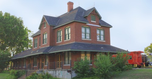 Historic brick train station with a gabled roof, large windows, and a red train car nearby, surrounded by greenery.