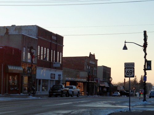 A quiet street scene at sunset, featuring small shops and a traffic sign for South 69, with snow on the ground.