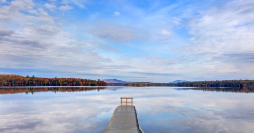 A serene lake scene with a wooden dock extending into calm waters, surrounded by colorful autumn foliage and a blue sky.
