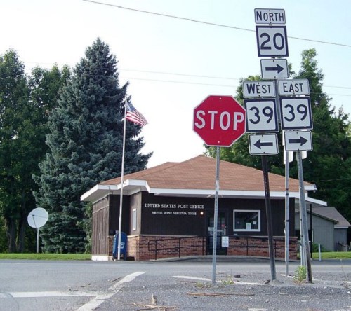 A small United States Post Office building with road signs indicating directions and a stop sign in front.