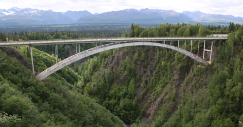 A large arch bridge spans a deep canyon, surrounded by lush greenery and mountains in the background.