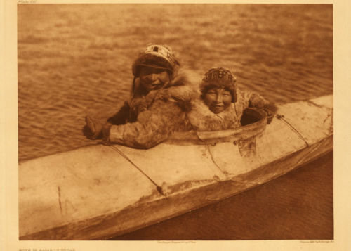 Two smiling children in a kayak, wearing fur clothing, against a backdrop of water.