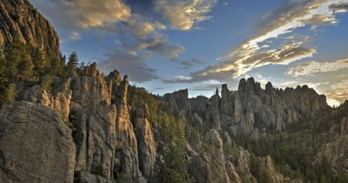 A dramatic landscape of rugged rock formations and trees under a colorful sky at sunset.