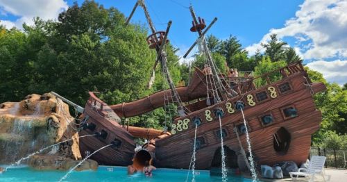 A large pirate ship water slide in a pool, surrounded by trees and featuring water sprays and a rocky structure.