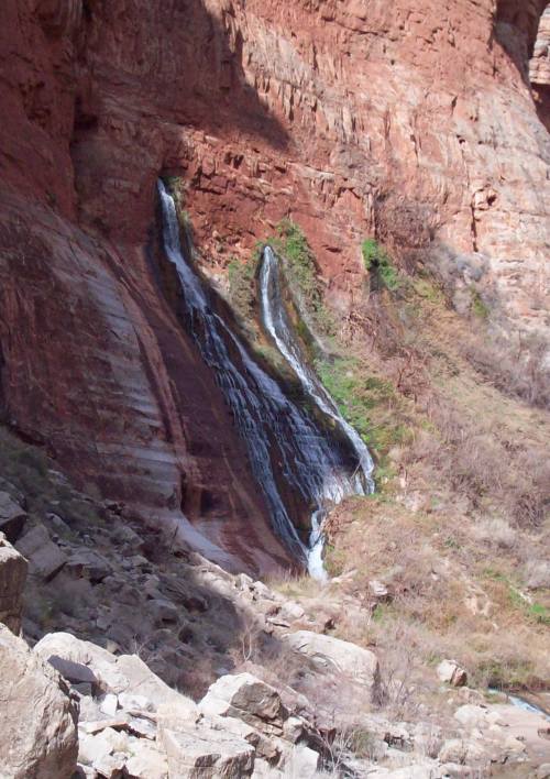 A waterfall cascading down red rock formations, surrounded by greenery and rocky terrain.