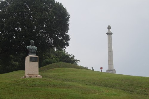 A bust and a tall monument stand on a grassy hill under a cloudy sky.