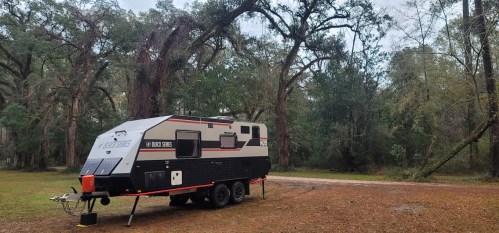 A compact camper trailer parked in a wooded area with tall trees and a grassy ground.