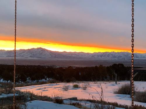 A vibrant sunset casts orange and yellow hues over snow-covered mountains, framed by trees and a snowy foreground.