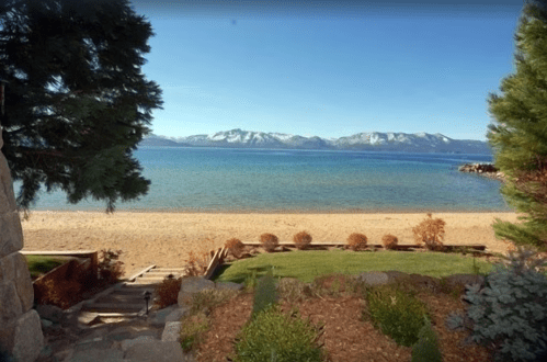 Scenic view of a sandy beach by a lake, framed by trees and mountains in the background under a clear blue sky.