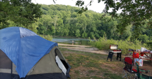 A blue tent by a river, surrounded by trees, with a camping setup including a grill and chairs nearby.