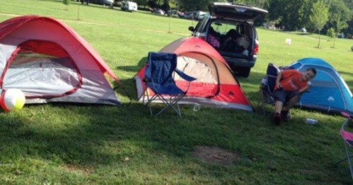 Two tents and a camping chair are set up on grass, with a person sitting nearby and a car in the background.