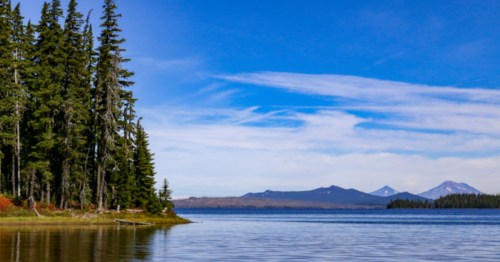 A serene lake surrounded by tall trees, with distant mountains under a clear blue sky.
