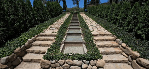 A serene garden pathway with stone steps and a water feature, flanked by lush greenery and trees under a clear blue sky.