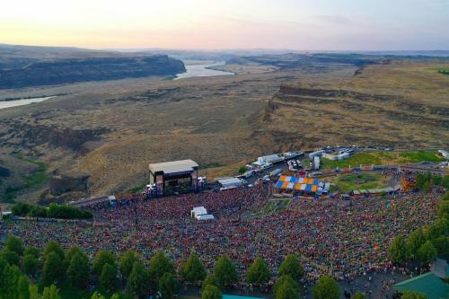 Aerial view of a large outdoor concert with a crowd, surrounded by hills and a river in the background at sunset.