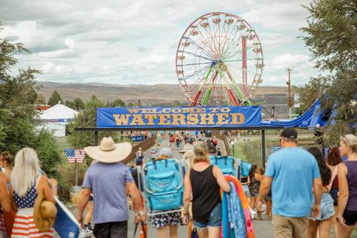 A crowd walks under a "Welcome to Watershed" sign, with a ferris wheel in the background and festive decorations.