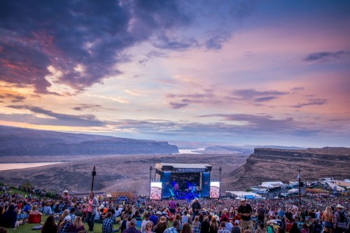 A large crowd gathers on a hillside at a music festival, with a scenic river and mountains under a colorful sunset.