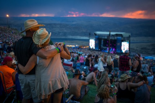 A couple embraces at a music festival, surrounded by a large crowd and a vibrant sunset in the background.