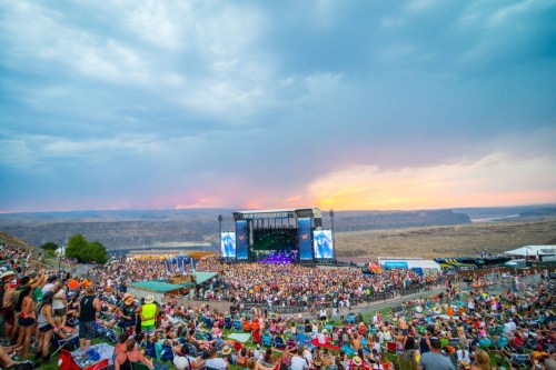 A large crowd enjoys a concert at a scenic outdoor venue during sunset, with mountains and a river in the background.