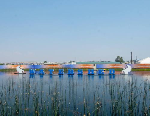 A calm water body with a sign reading "WATERSHED" and greenery in the foreground under a clear blue sky.