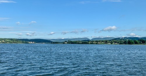 A serene lake view with gentle waves, surrounded by green hills and a clear blue sky dotted with clouds.