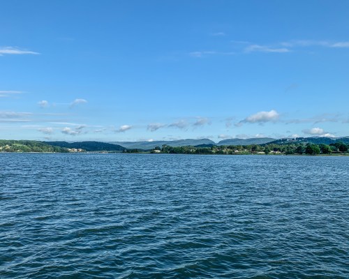 A serene lake view with gentle waves, surrounded by green hills and a clear blue sky with scattered clouds.
