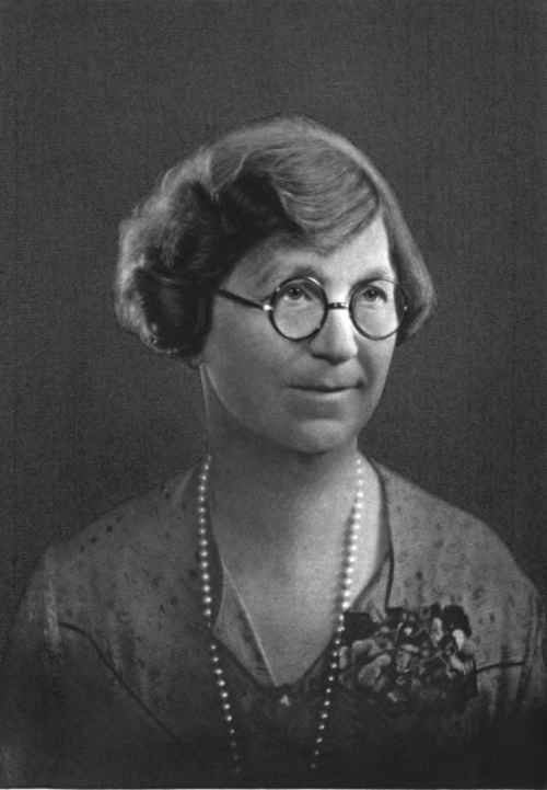Black and white portrait of a woman with glasses, short hair, and a pearl necklace, smiling softly at the camera.