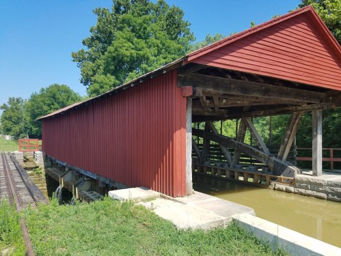 A red covered bridge beside a calm waterway, surrounded by green trees and a clear blue sky.