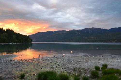 A serene lake at sunset, reflecting colorful clouds and surrounded by mountains and trees.