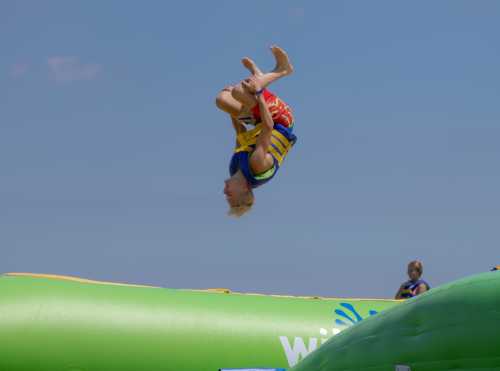 A child performs a backflip in mid-air above a green inflatable at a beach on a sunny day.