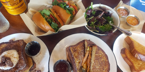 A table spread featuring sandwiches, a salad, sweet potato fries, and syrup in small cups.