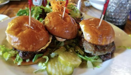Three mini burgers on a plate, topped with lettuce, pickles, and a side of fresh salad.