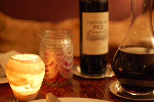A softly lit table setting featuring a candle, a decorative jar, a bottle of wine, and a wine decanter.