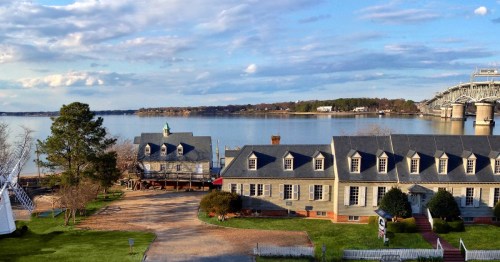 Scenic view of a waterfront with historic buildings, trees, and a bridge under a partly cloudy sky.