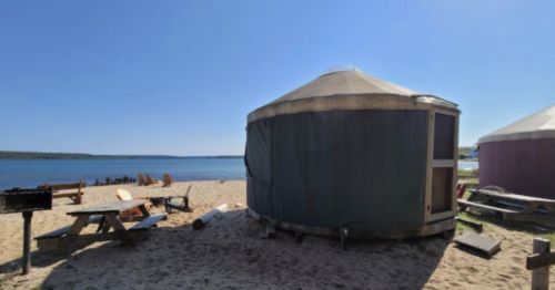 A yurt on a sandy beach with a view of the water, surrounded by picnic tables and chairs under a clear blue sky.