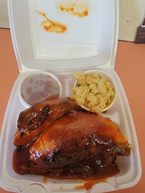 A takeout container with barbecue chicken, macaroni and cheese, and a side of sauce.