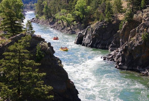 A river flows through rocky cliffs, with a group rafting in orange boats amidst lush green trees.